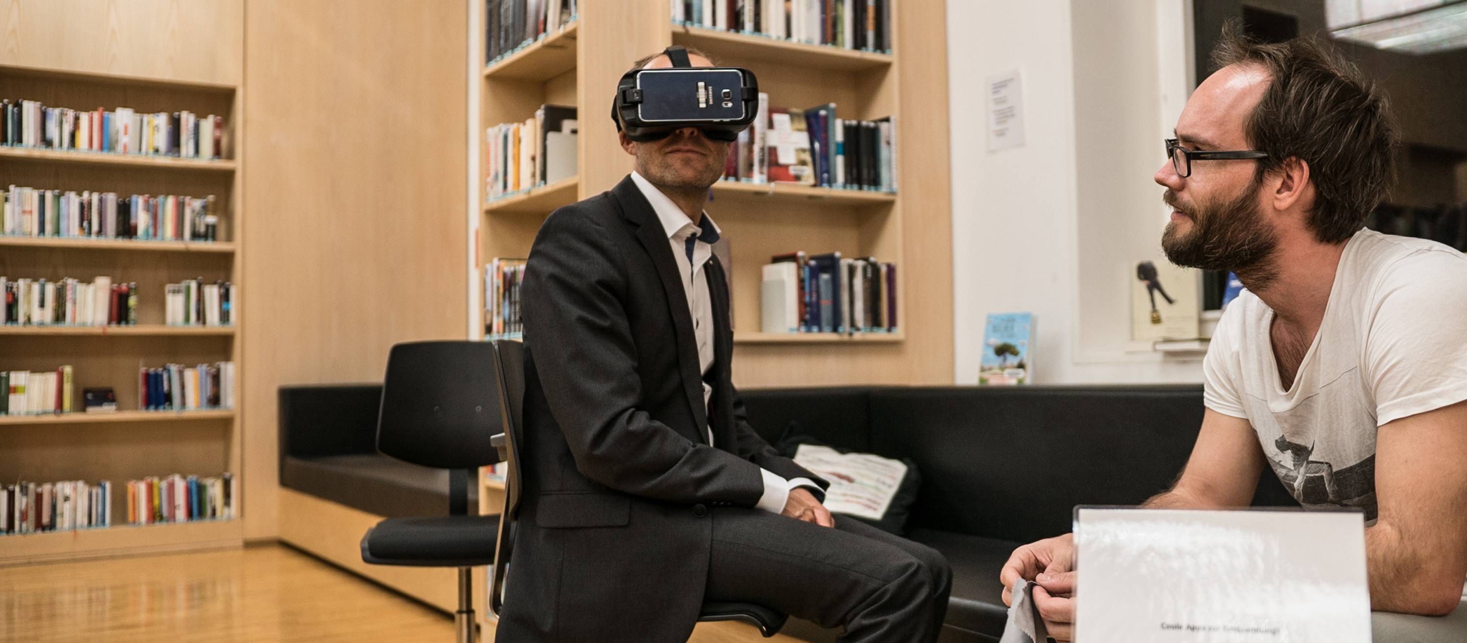 Blick auf zwei Personen sitzen bei einer Veranstaltung in der Bibliothek, einer trägt eine Virtual Reality Brille