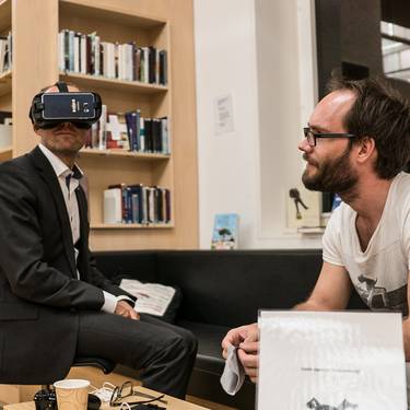 Blick auf zwei Personen sitzen bei einer Veranstaltung in der Bibliothek, einer trägt eine Virtual Reality Brille