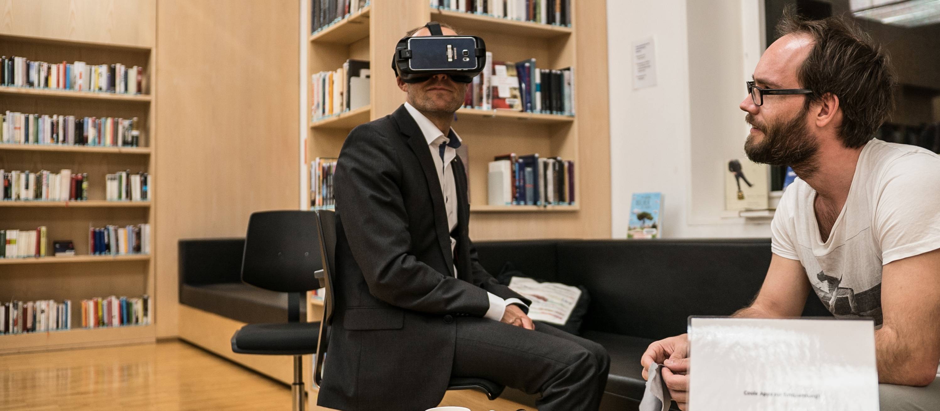 Blick auf zwei Personen sitzen bei einer Veranstaltung in der Bibliothek, einer trägt eine Virtual Reality Brille