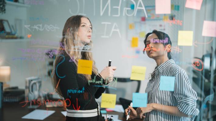 Fundiert und aktuell Zwei Frauen arbeiten an einer Glaswand mit Haftnotizen und entwickeln gemeinsam Ideen.