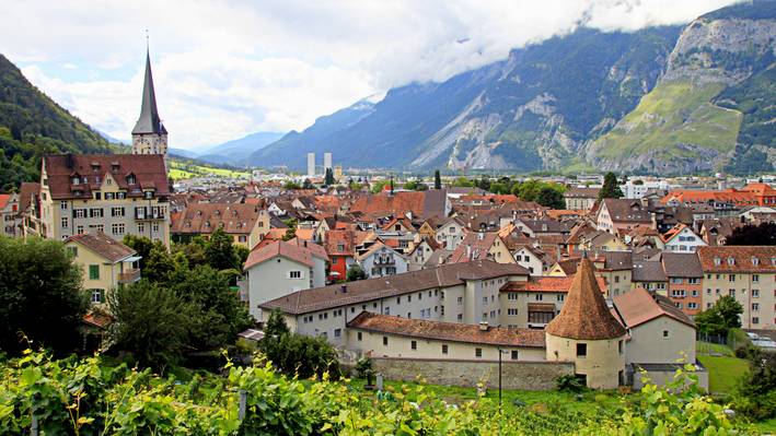Blick über die Altstadt von Chur mit Kirchturm, Weinreben und Bergkulisse im Hintergrund.