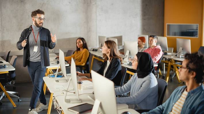 Lehrer unterrichtet und läuft durchs Klassenzimmer, Studierende sitzen am PC und hören ihm zu.