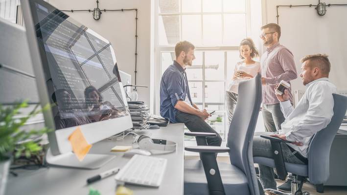 Vier Personen sitzen und stehen beisammen und diskutieren miteinander, im Vordergrund ist ein Arbeitsplatz mit Bildschirm zu sehen.