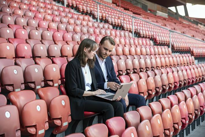 Sportwissenschaft Studium: Zwei Studierende sitzen mit dem Laptop im Stadion.