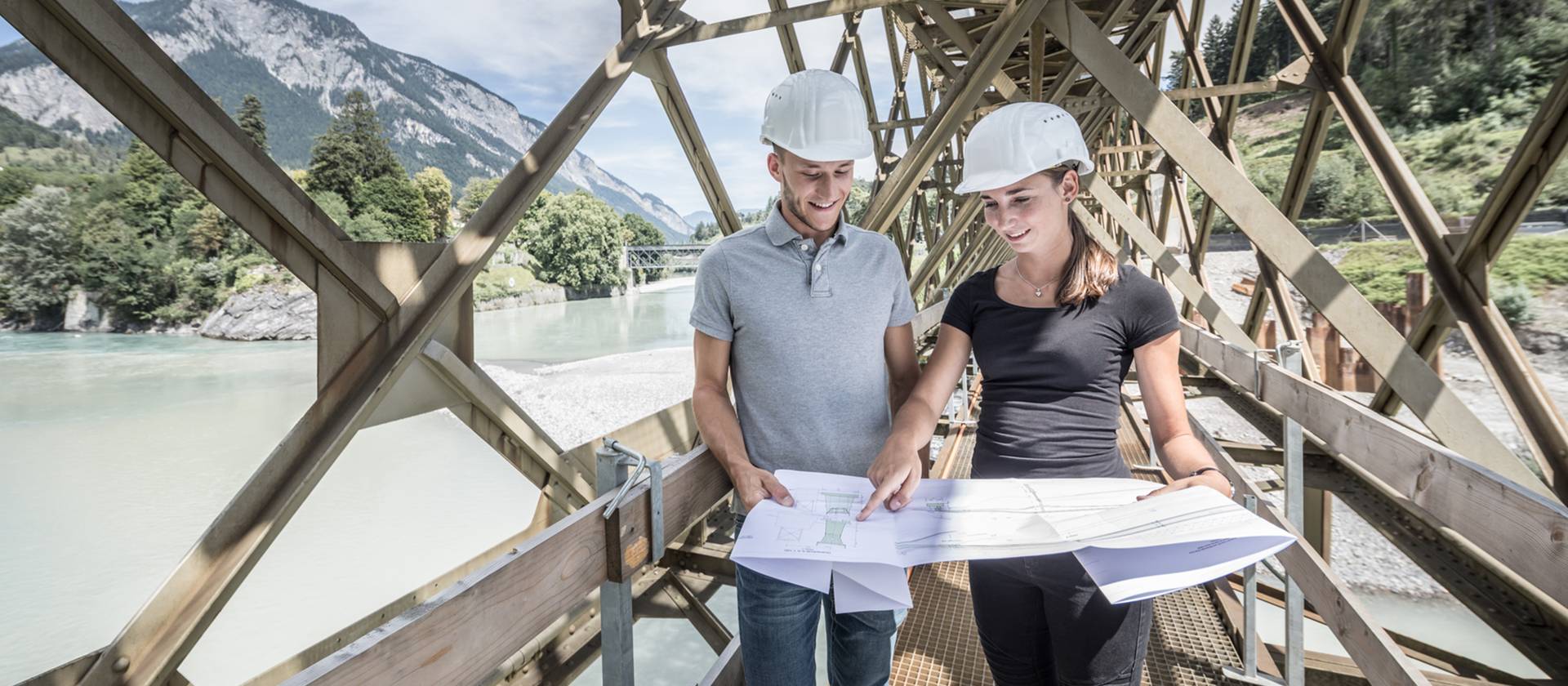 Angehende Bauingenieurin und Bauingenieur der Fachhochschule Graubünden mit Plänen in den Händen auf einer Brücke.
