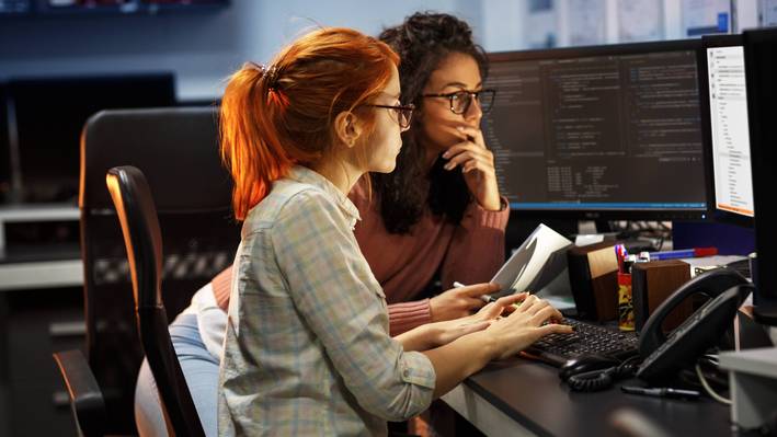 Eine Frau sitzt vor dem Computer, eine zweite steht daneben und zusammen probieren sie ein Problem auf dem Bildschirm zu lösen. 