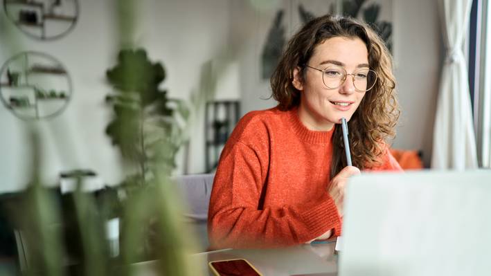 A smiling student following a webinar on her laptop at home and taking notes