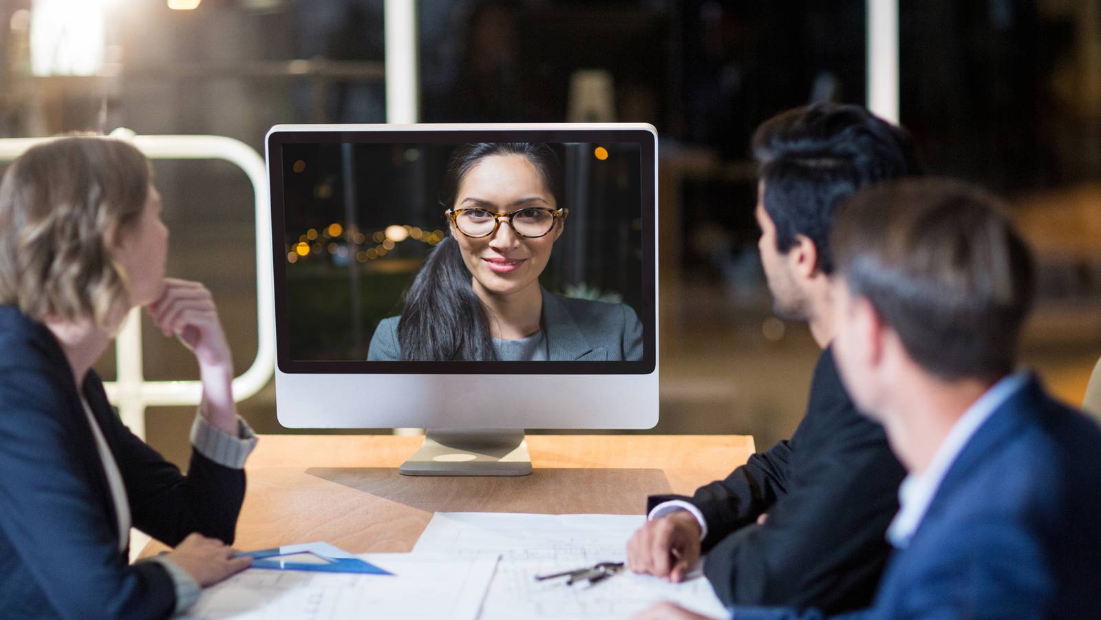 Personen an einem Konferenztisch, die auf einen Monitor blicken in dem eine Frau virtuell an der Sitzung teilnimmt
