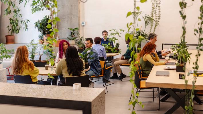Gruppe von Studierenden im Gespräch an Tischen in einem begrünten Coworking-Raum