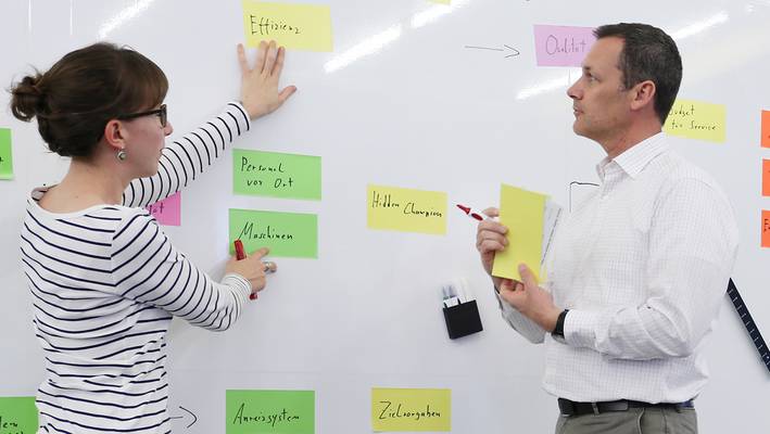 Eine Frau und ein Mann, die vor einem Whiteboard stehen und die darauf gehefteten Zettel diskutieren