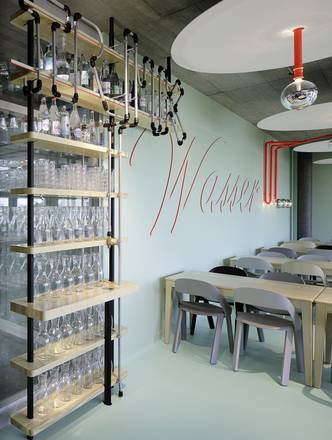 Cafeteria mit Regal aus Rohrelementen und Flaschen, Schriftzug «Wasser» an mintgrüner Wand, Tische und Stühle im Hintergrund.